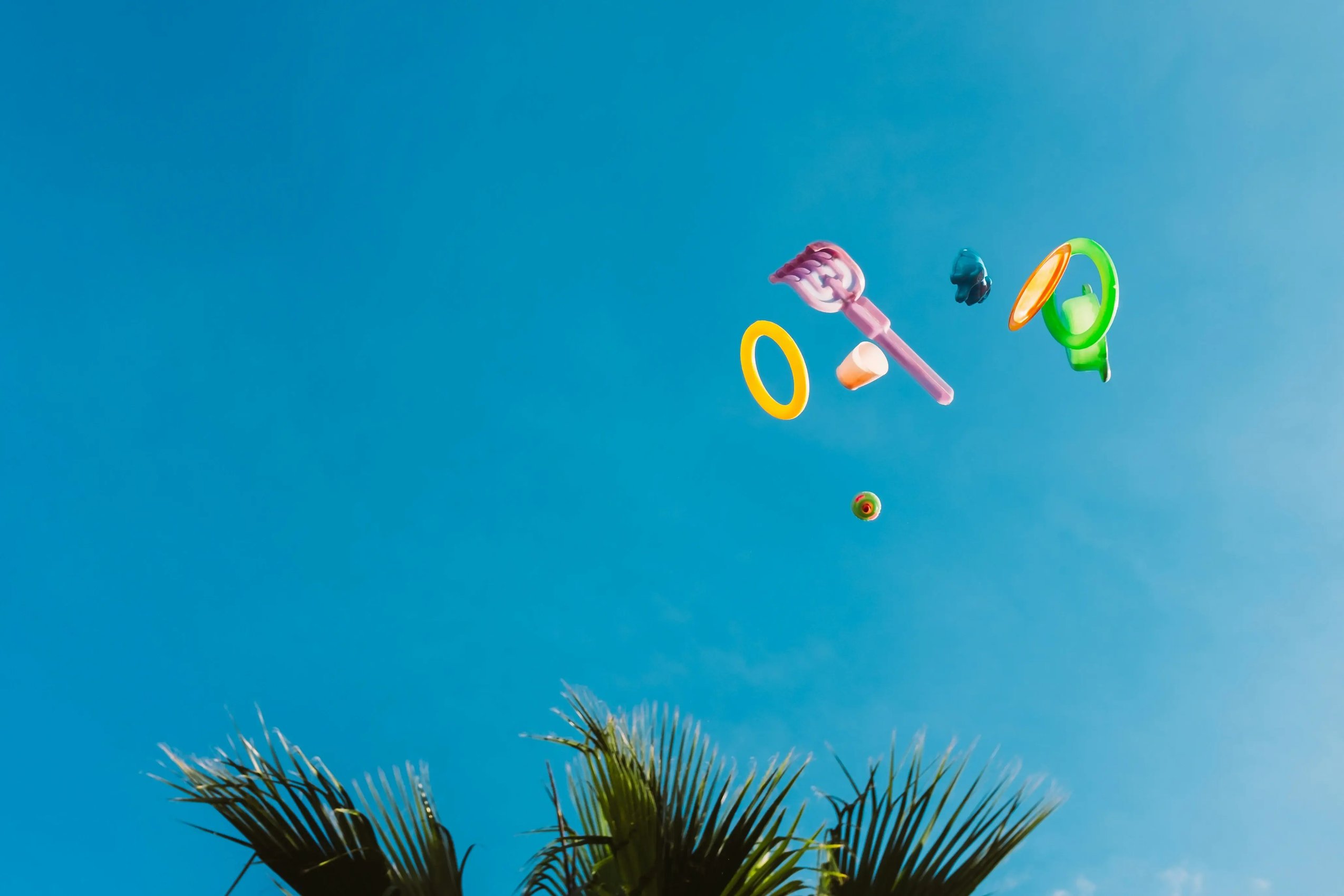 Various colorful toys floating against a clear blue sky with palm fronds at the bottom, reflecting the expansive and diverse applications of play in real-world settings.
